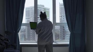 Rear view of woman in pajamas at home standing by the window and typing. Media. Young woman working on laptop with green screen. photo