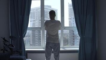 Rear view of a pensive man standing at home by the window and looking at the city. Media. Man in home clothes spending time in reflection in his bedroom. photo