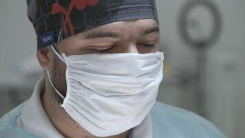 de gezicht van een mannetje chirurg in een steriel masker in in werking kamer. actie. dichtbij omhoog van een dokter gedurende chirurgie. video