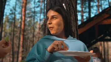 hermosa morena mujer en pie por el árbol y comiendo. valores imágenes. mujer tomando comida desde lámina, sonriente en el antecedentes de bosque y cielo. video