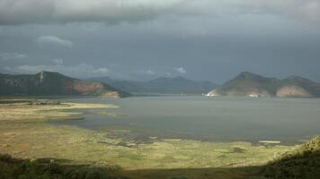 Sun and shadow on green meadow and a lake. Creative. Calm water and mountains on cloudy sky background. video