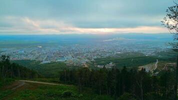 Aerial flight downhill the green mountains towards tourist town. Clip. Summer green trees on a hill slope and the city with cloudy sky above. video