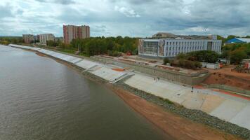 Aerial view of city embankment with construction site. Clip. Unfinished area and calm river with buildings and green trees on the background. video