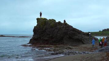 Man stands on rock on seashore. Clip. Group of people on seashore with rock on cloudy day. Sea rock with group of tourists on coast video