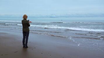 donna su riva prende immagini di mare su Telefono. clip. donna prende immagini di mare su nuvoloso giorno. donna prende immagini di mare con onde video