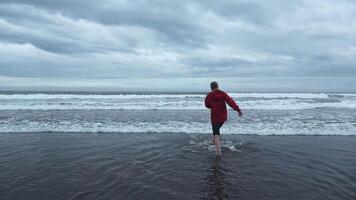 Mann Spaziergänge im Wasser auf Ufer mit Wellen. Clip. Mann Spaziergänge im Wasser auf Hintergrund von Meer Horizont auf wolkig Tag. Mann benetzt seine Füße im Meer auf wolkig Tag video