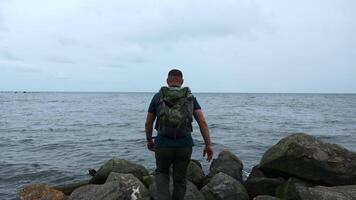 Man walks on stone coast on background of sea. Clip. Man walks on stones of sea embankment on cloudy day. Walk along sea stone coast on cloudy day video