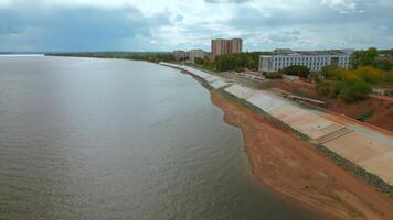 Aerial view of city embankment with construction site. Clip. Unfinished area and calm river with buildings and green trees on the background. video