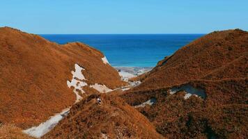 Couple walking along path on hills on background of sea. Clip. Top view of hills with trails and autumn grass on background of sea coast. Beautiful autumn hills by sea on sunny day video