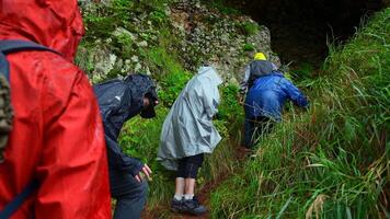 grupo de turistas Vamos a cueva. acortar. turistas entrar cueva en rocas en lluvioso día. grupo de personas en impermeables en rocoso montañas Vamos a cueva en verano video