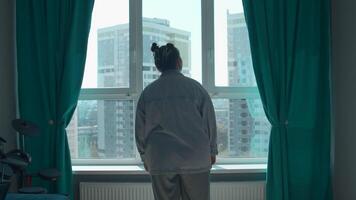 ung kvinna njuter se genom de panorama- fönster i de morgon- på Hem. media. flicka i pyjamas stående och tänkande förbi de fönster i sovrum. video