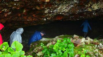 Group of tourists go to cave. Clip. Tourists enter cave in rocks on rainy day. Group of people in raincoats in rocky Mountains go to cave in summer video