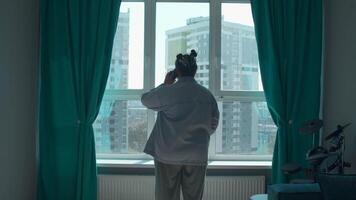 Rear view of a woman talking on a cell phone by the window. Media. Woman in pajamas talking on smartphone at home. video