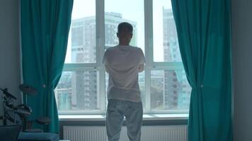 Rear view of a pensive man standing at home by the window and looking at the city. Media. Man in home clothes spending time in reflection in his bedroom. video