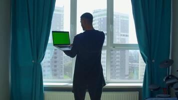 Rückseite Aussicht von ein männlich Schüler studieren beim Zuhause und mit seine Laptop. Medien. Mann Stehen durch das Fenster und suchen beim Grün Chroma Schlüssel Bildschirm. video
