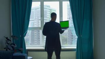 Rear view of a male student studying at home and using his laptop. Media. Man standing by the window and looking at green chroma key screen. video