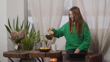 Young woman in glasses having break from work at home. Media. Red haired lady at kitchen drinking tea and watching movie on a tablet. video