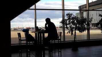 homem de negocios esperando para embarque às a aeroporto e olhando através panorâmico janelas. silhueta do uma homem às uma mesa. video