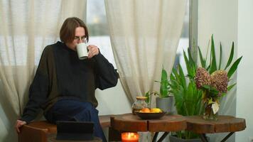 Handsome student drinks tea and looks at tablet in cafe. Media. Young man with glasses is watching lecture on tablet in cafe. Young man is relaxing and studying in cozy cafe video