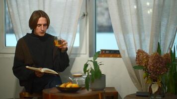 Young handsome man is reading book with tea in cafe. Media. Student is sitting alone and reading book in cozy cafe. Young man is reading book and drinking tea in student cafe video