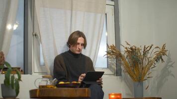 Young man drinks tea and works on tablet in cafe. Media. Student is relaxing with tablet and tea in cozy cafe. Handsome student studying on break in cafe video