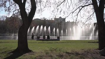 City fountain. Footage. People walking in the park. Fountain in city park on hot summer day. Stream of water, drops and bright splashes of water in beautiful city fountain video