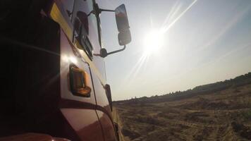 Side view of red truck cab moving on dusty unpaved rural road along green trees ahead. Scene. Huge lorry riding on country, dusty road on cloudy sky background, view from a wheel. video