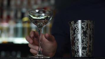 Close up of a bartender putting ice cubes into the small empty cocktail glass. Media. Details of making a drink with blurred bottles of alcohol on the background. video