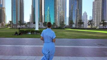 Rear view of a young woman running on stone path in a city park, Dubai. Action. Woman spending time outdoors near tall buildings. video