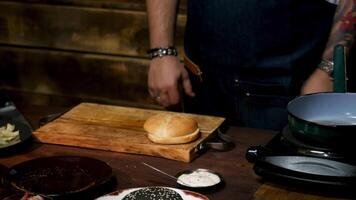 cerca arriba de masculino cocinero manos tomando hamburguesa bollo y poniendo eso en un de madera tablero rodeado por diferente utensilios de cocina y ingredientes. valores imágenes. proceso de delicioso hamburguesa preparación. video