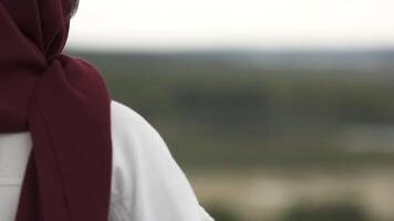 posteriore Visualizza di un' donna in piedi sopra il verde foresta, campo, e fiume. azione filmato. mutevole il messa a fuoco a partire dal il femmina vestito nel rosso sciarpa su sua testa e bianca giacca per il naturale primavera o video