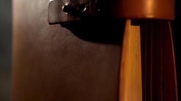 Vintage leather book with yellow pages. Stock footage. Close up of an ancient notebook with brown leather cover and a strap in vertical position isolated on black background under the dim light. video