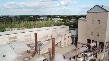 Aerial view of a small factory with smoking chmneys. Stock footage. Flying above the factory surrounded by green trees on blue cloudy sky background, industrial works concept. video