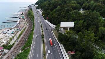 aereo Visualizza di il strada con in movimento macchine e un' camion lungo il montagna pendenza coperto di verde alberi su uno lato e il mare su il altro lato. scena. superiore Visualizza di ferrovia e automobile strada con video