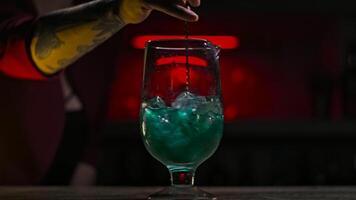 Close-up of bartender stirring cocktail with ice with spoon. Stock footage. Beautiful mixing with cocktail spoon of colored drink with ice on dark neon background video