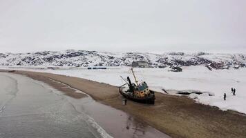 abbandonato vecchio nave su un' sabbioso riva di il mare con neve coperto colline. filmato. aereo Visualizza di bellissimo inverno paesaggio con un' distrutto nave, persone a piedi su il riva e il mare. video