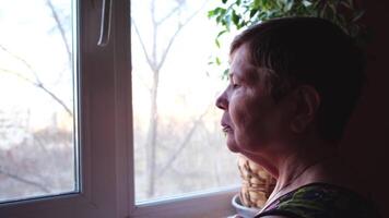 ett äldre kvinna ser tyvärr genom hela de fönster. begrepp. sida se av fundersam omtänksam ensam senior vuxen mormor stående ensam på Hem ser genom fönster och väntar för någon. video
