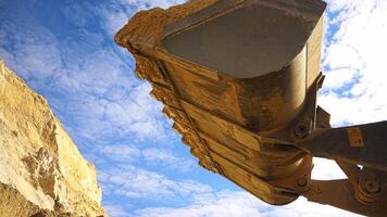 Excavator in action at the construction site. Stock footage. Bottom view of excavator bucket of yellow color loaded with sand on blue cloudy sky background. video