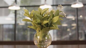 Gentle lilly of valley flowers in glass vase standing on wooden table on blurred background with windows and lamps. Stock footage. Spring may lily bouquet, decoration of room. video