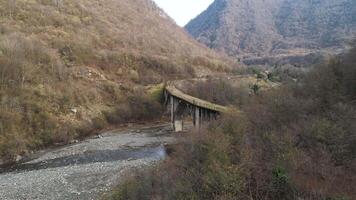 lado ver de un antiguo abandonado puente en un antecedentes de montañas en el pequeño pueblo en tarde otoño estación. disparo. aéreo de cubierto de musgo Roca puente y el estrecho arroyo. video