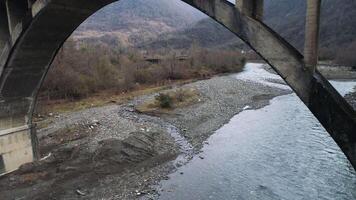 vliegend onder roestig brug kruispunt de versmallen rivier- met steenachtig bodem omringd door bergen en Woud. schot. antenne van landelijk landschap in laat herfst. video