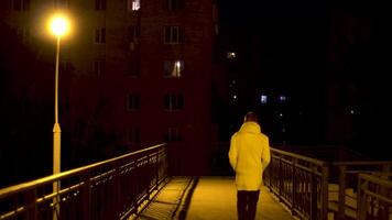 peatonal puente con un solitario calle lámpara y un hombre cruce eso a noche. valores imágenes. posterior ver de un joven hombre en blanco chaqueta cruce el puente en el ciudad a noche. video