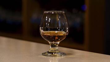 Close up of a glass with cognac, brandy or whisky standing on the bar counter. Stock footage. Strong strong alcoholic beverage inside a glass on the blurred background of a dark room. video