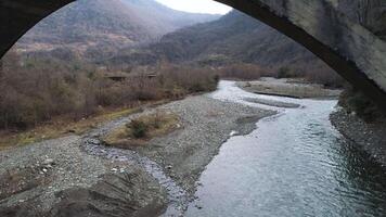 Flying under the destroyed, broken concrete bridge against the background of autumn forest and a river with stony shore. Shot. Aerial of cold mountainous stream and forested hills. video