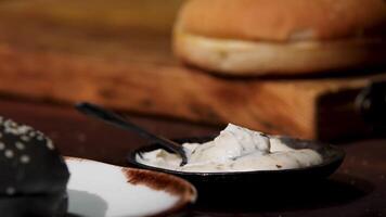 Close up of white sauce in black bowl standing on the wooden table near black and white burger buns, food ingredients concept. Stock footage. Home made mayonnaise sauce. video