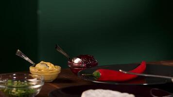 Close up of different ingredients for cooking standing on a wooden table on green wall background. Stock foorage. Chili pepper, cranberry, mayonnaise, and mustard sauces, camembert cheese and greenery video