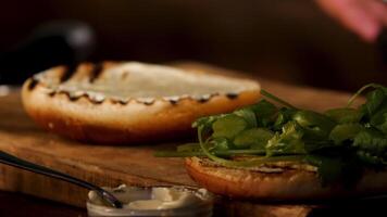 Craft burger is being cooked on dark room background. Stock footage. Close up of white sour cream sauce on a toasted burger bun lying on a wooden board near greenery. video