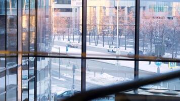 se från inuti de företag byggnad zon genom de glas fönster på en stad gata med rör på sig bilar. stock antal fot. vinter- väg med körning bilar och panorama- fönster av företag Centrum. video