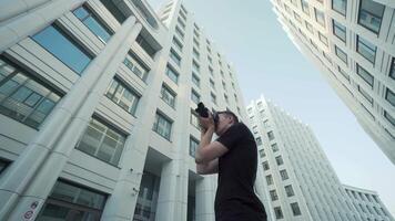 Young photographer takes picture of modern urban architecture. Action. Bottom view of guy photographing modern architecture with camera. Photographer shoots office buildings against blue sky video