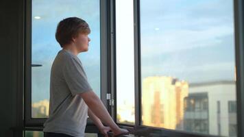 Jeune homme jouit vue de fenêtre permanent sur balcon. concept. étudiant est Heureusement en train de regarder la vie à l'extérieur fenêtre sur Contexte ciel. étudiant des stands sur balcon et regards en dehors fenêtre video